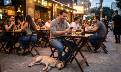 Homem sorri olhando celular em bar lotado com pessoas e cachorro dormindo no chão