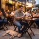 Homem sorri olhando celular em bar lotado com pessoas e cachorro dormindo no chão