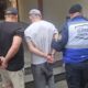 Agentes do Segurança Presente escoltam dois homens algemados, um deles usando colete com 'SEGURANÇA PRESENTE'