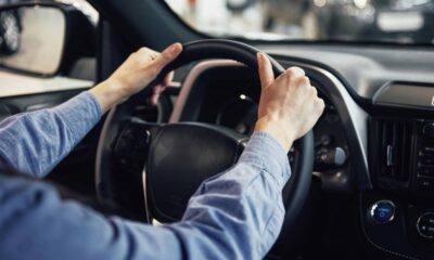 Mãos de pessoa em camisa azul segurando volante de carro com painel e botões AUTO OFF