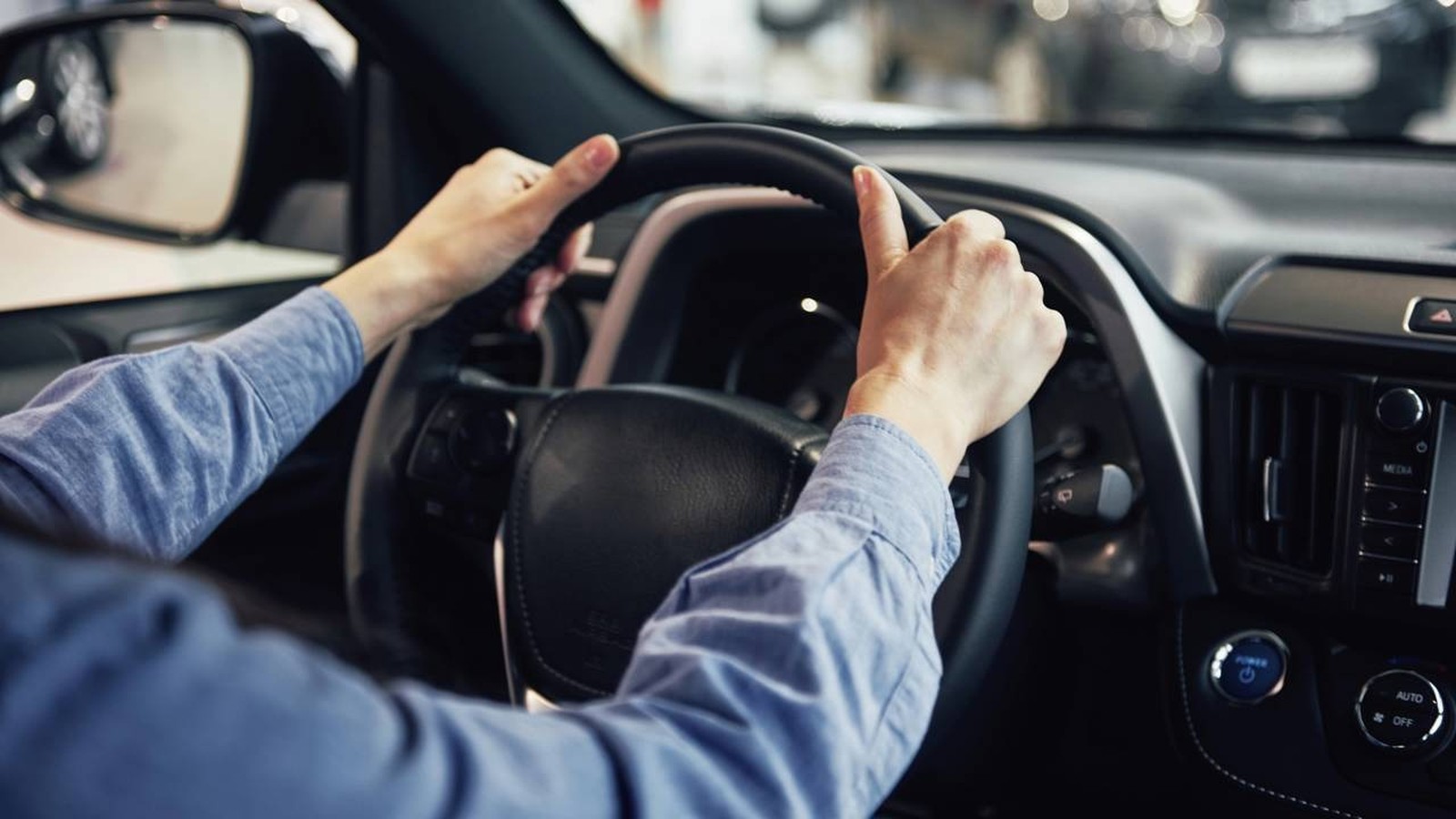 Mãos de pessoa em camisa azul segurando volante de carro com painel e botões AUTO OFF