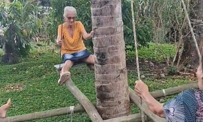 Mulher idosa sentada em balanço de madeira, segurando tronco de árvore, em um quintal