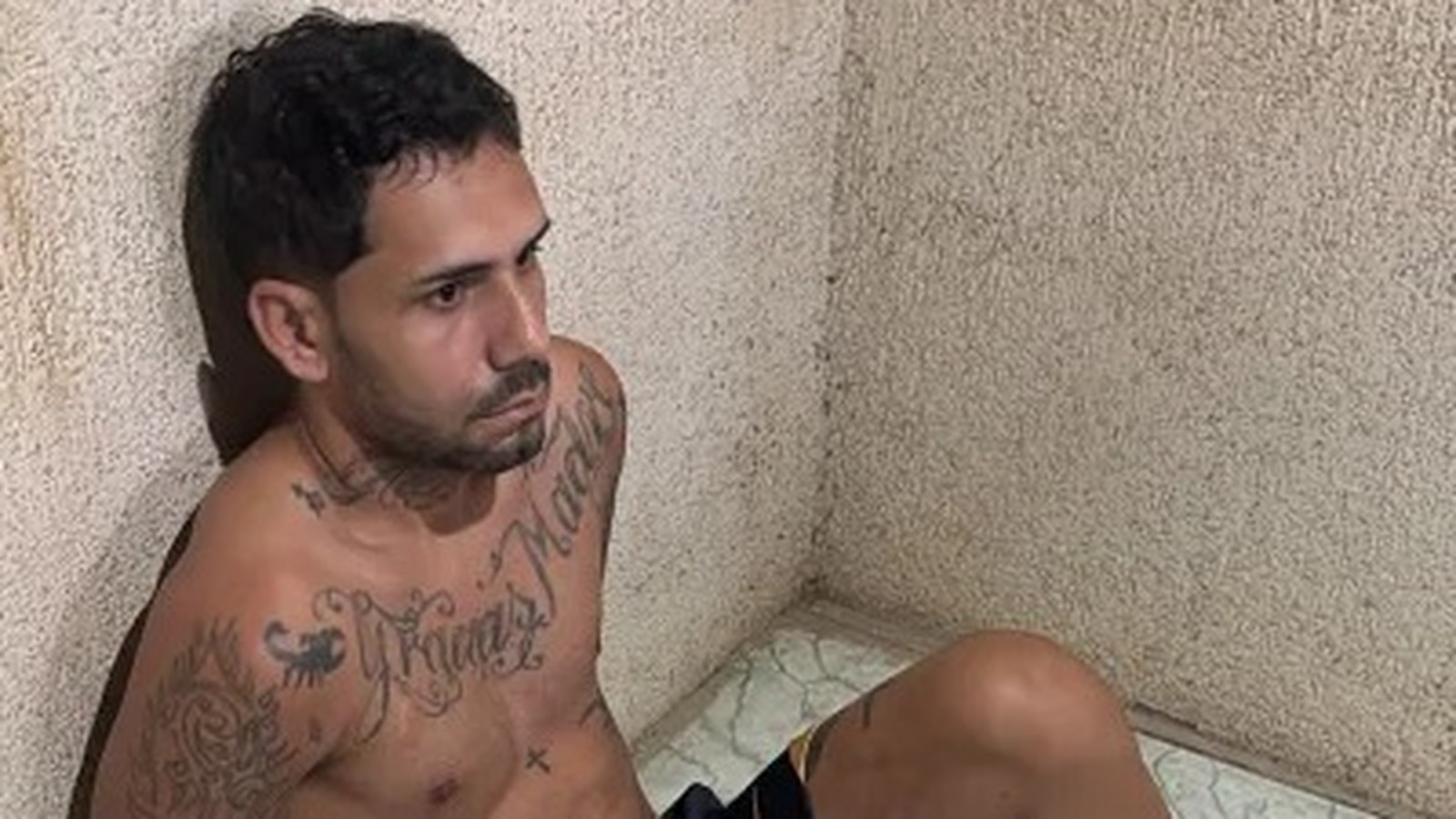 Homem tatuado, sem camisa, com barba e cabelo escuro, sentado contra parede de textura clara