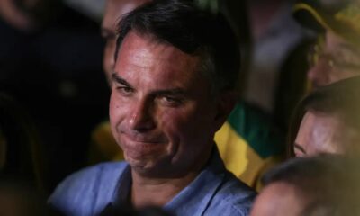 Close-up de Flávio Bolsonaro, cabelo escuro, camisa azul, rosto sério, olhando para o lado.