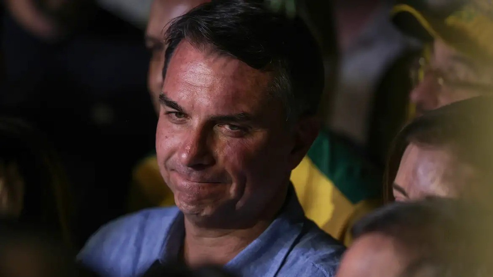Close-up de Flávio Bolsonaro, cabelo escuro, camisa azul, rosto sério, olhando para o lado.