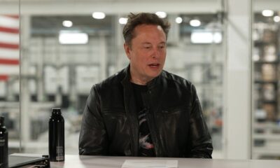 Elon Musk sentado em mesa branca, jaqueta preta, com duas garrafas escuras, fundo industrial