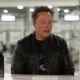 Elon Musk sentado em mesa branca, jaqueta preta, com duas garrafas escuras, fundo industrial