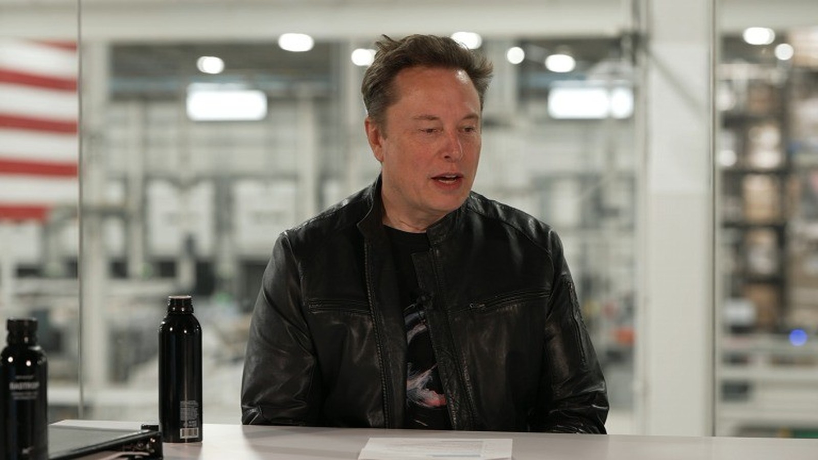 Elon Musk sentado em mesa branca, jaqueta preta, com duas garrafas escuras, fundo industrial
