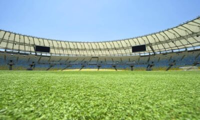 Gramado de futebol verde dentro de estádio vazio com assentos azuis e amarelos e telões