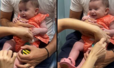 Bebê sorridente em vestido laranja tomando vacina no colo, com mãos de profissionais.