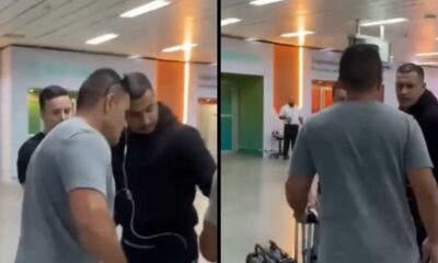 Homem de camiseta cinza escoltado por dois homens em preto no saguão do aeroporto