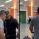 Homem de camiseta cinza escoltado por dois homens em preto no saguão do aeroporto