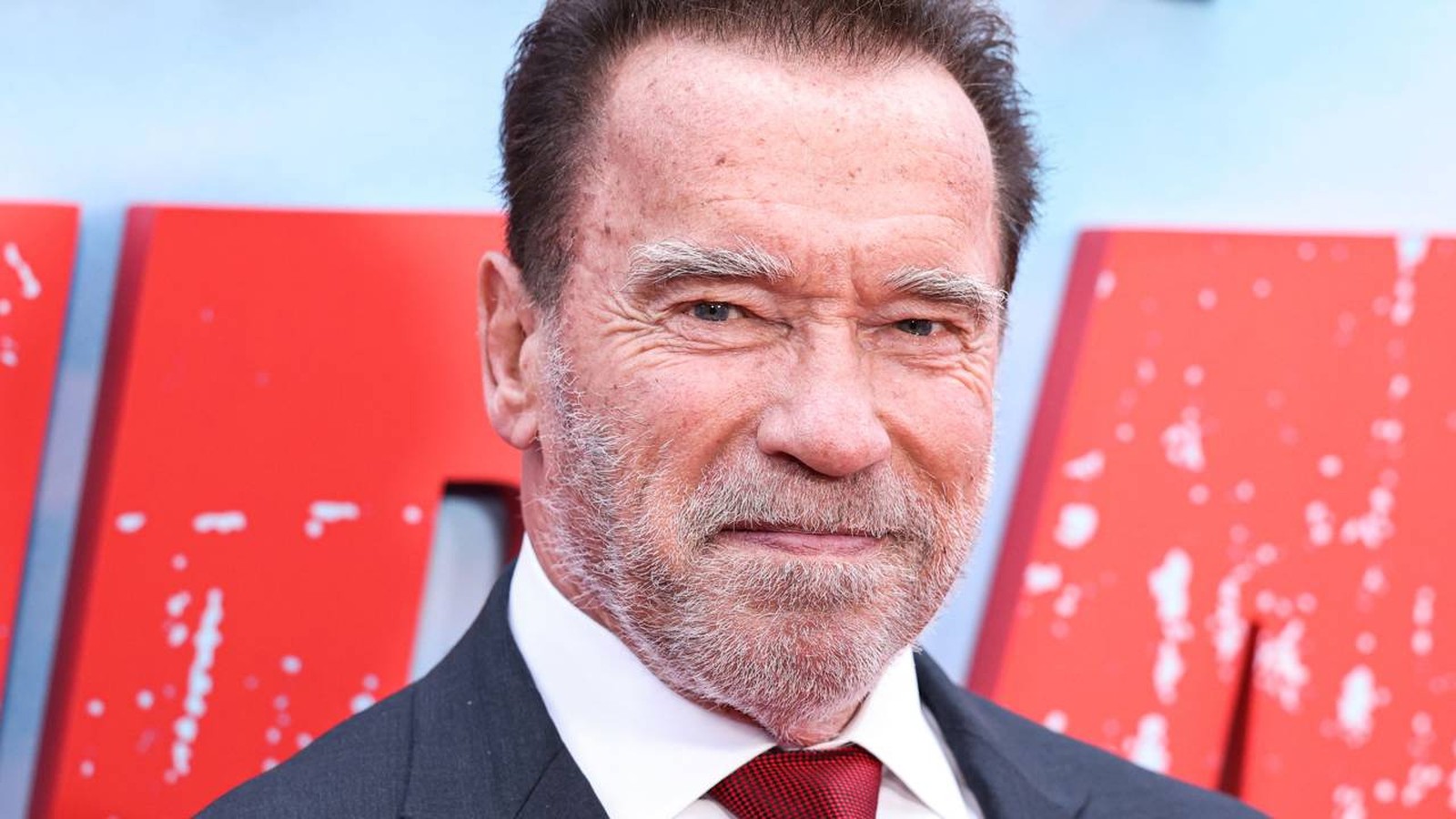 Rosto de Arnold Schwarzenegger em close, sorrindo e usando terno e gravata vermelha