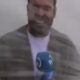 Homem com microfone azul e casaco cinza, expressão com boca aberta, em fundo claro borrado