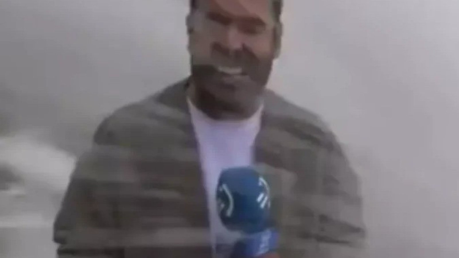 Homem com microfone azul e casaco cinza, expressão com boca aberta, em fundo claro borrado