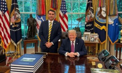 Donald Trump e Flávio Bolsonaro sorrindo no Salão Oval da Casa Branca com bandeiras ao fundo
