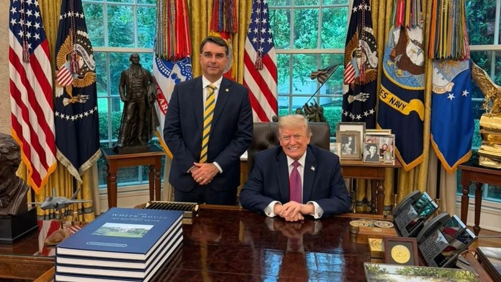 Donald Trump e Flávio Bolsonaro sorrindo no Salão Oval da Casa Branca com bandeiras ao fundo