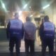 Três homens vistos de costas, dois usando coletes 'Segurança Presente' escoltam um homem com mãos para trás