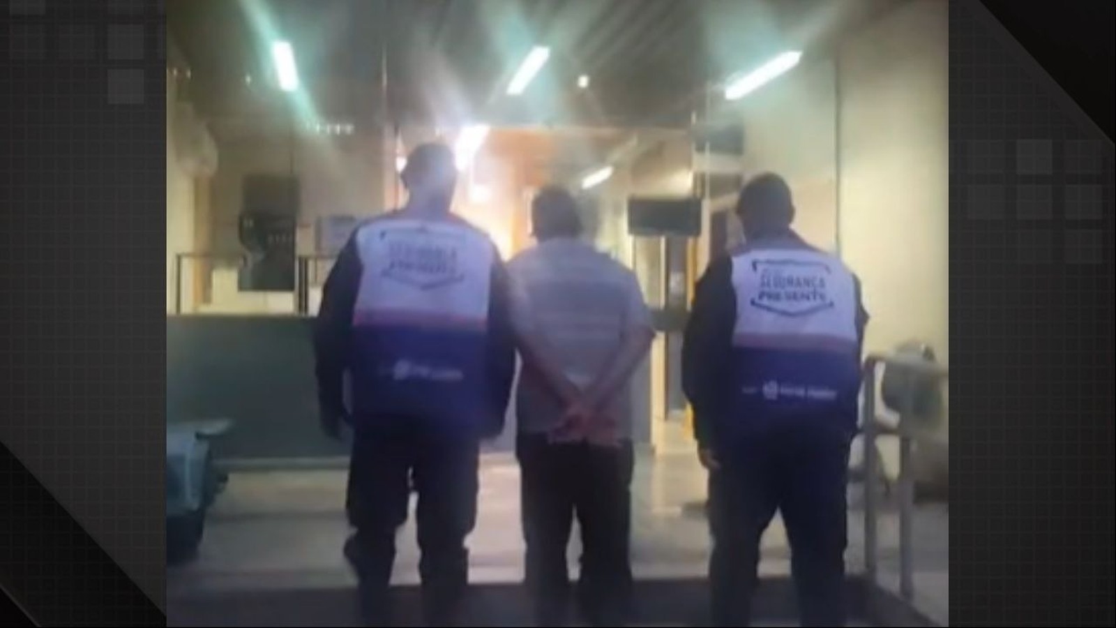 Três homens vistos de costas, dois usando coletes 'Segurança Presente' escoltam um homem com mãos para trás