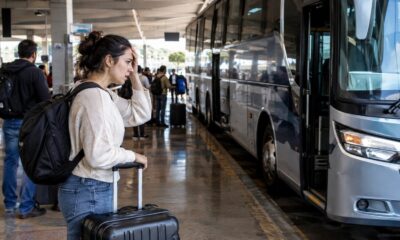 Mulher com mochila, mão na testa, e mala em estação rodoviária com ônibus.