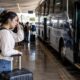 Mulher com mochila, mão na testa, e mala em estação rodoviária com ônibus.