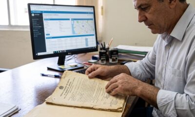 Homem examinando certidão de registro de imóvel antiga, com computador exibindo sistema digital