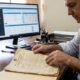 Homem examinando certidão de registro de imóvel antiga, com computador exibindo sistema digital