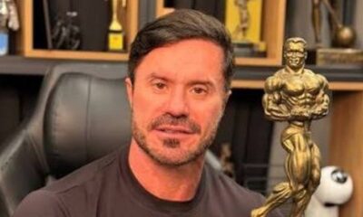 Renato Cariani de camiseta preta e barba segura um troféu dourado de fisiculturismo, em ambiente interno