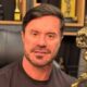 Renato Cariani de camiseta preta e barba segura um troféu dourado de fisiculturismo, em ambiente interno