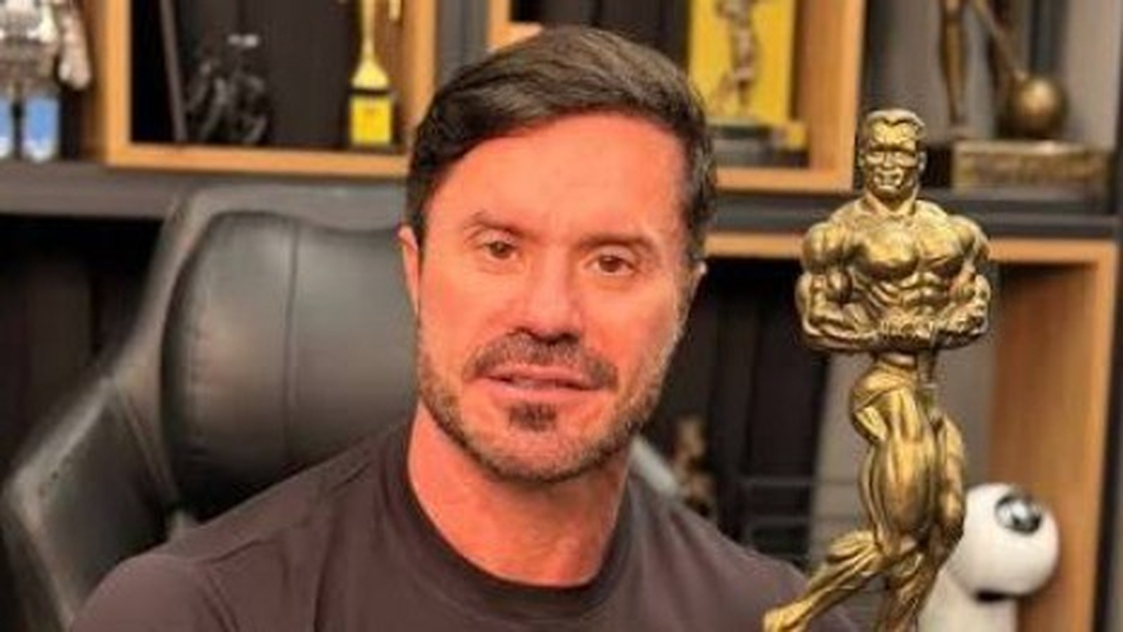 Renato Cariani de camiseta preta e barba segura um troféu dourado de fisiculturismo, em ambiente interno
