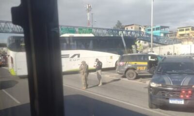 Ônibus branco e duas viaturas da PRF parados em rodovia, com ponte e prédios ao fundo