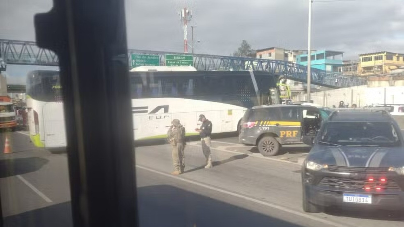 Ônibus branco e duas viaturas da PRF parados em rodovia, com ponte e prédios ao fundo