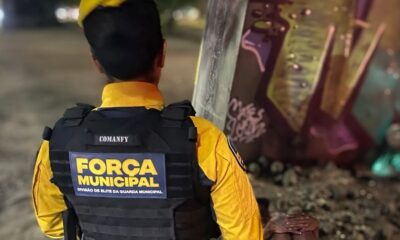Agente de costas em uniforme amarelo e colete tático preto com "FORÇA MUNICIPAL" e "COMANFY"