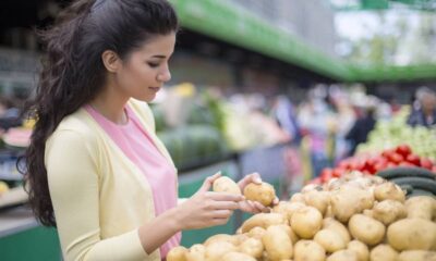 Mulher jovem de cabelos escuros inspeciona duas batatas em uma barraca de feira livre com pilhas de legumes