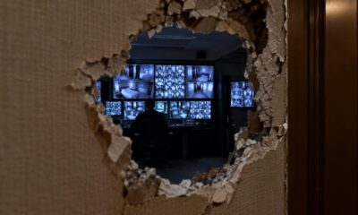 Buraco em parede revela pessoa em sala escura monitorando várias telas de vigilância com quartos