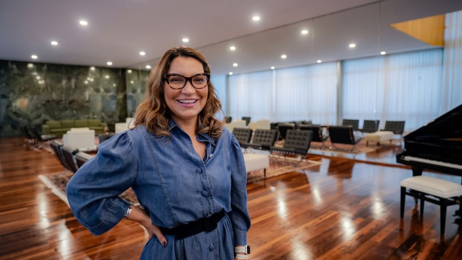 Janja da Silva, em pé e sorrindo, usa óculos em ambiente com piano e poltronas