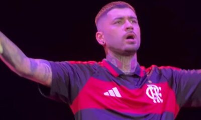 Zayn Malik em close-up, no palco, vestindo camisa preta e vermelha do Flamengo, com braços abertos