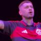 Zayn Malik em close-up, no palco, vestindo camisa preta e vermelha do Flamengo, com braços abertos