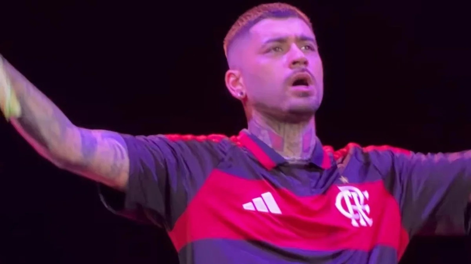 Zayn Malik em close-up, no palco, vestindo camisa preta e vermelha do Flamengo, com braços abertos