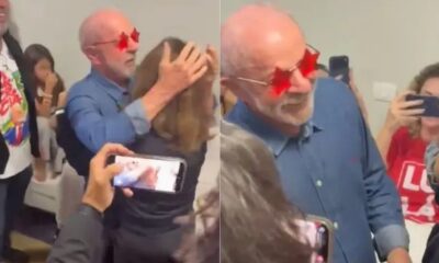 Lula, de óculos estrela, abraça mulher, pessoas filmam. Em segundo plano, camisa com "L"