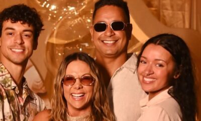 Quatro pessoas felizes, incluindo Carla Perez e Xanddy, sorrindo em um fundo dourado com balões