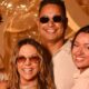 Quatro pessoas felizes, incluindo Carla Perez e Xanddy, sorrindo em um fundo dourado com balões