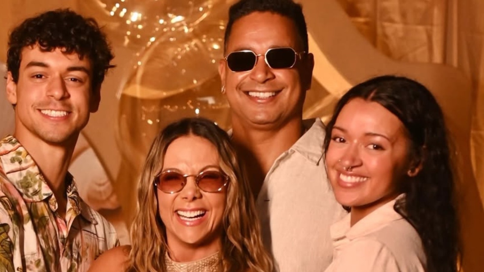 Quatro pessoas felizes, incluindo Carla Perez e Xanddy, sorrindo em um fundo dourado com balões