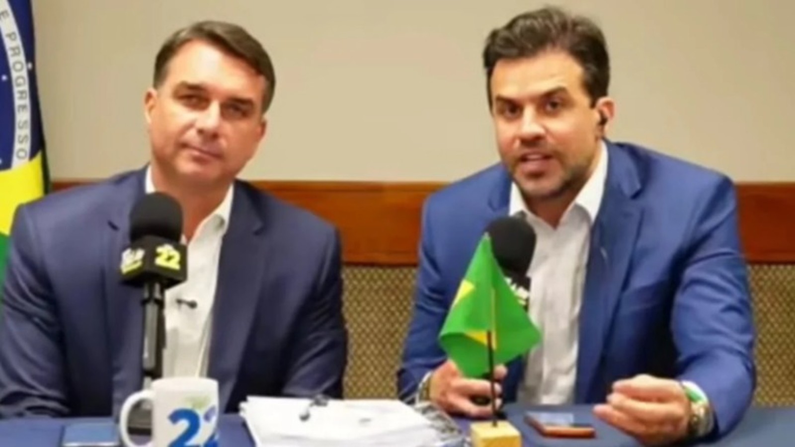 Flávio Bolsonaro e Pablo Marçal em ternos, sentados com microfones e bandeira do Brasil
