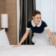 Mulher com uniforme de camareira arruma uma cama branca em um quarto de hotel
