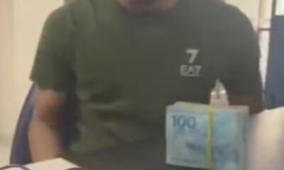 Homem de camiseta verde, logo 7 EA7. Maço de notas de R$ 100 na mesa.