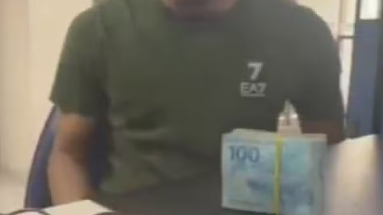 Homem de camiseta verde, logo 7 EA7. Maço de notas de R$ 100 na mesa.