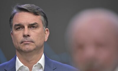 Flávio Bolsonaro, de terno, sério em primeiro plano, com figura borrada ao fundo