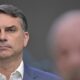 Flávio Bolsonaro, de terno, sério em primeiro plano, com figura borrada ao fundo