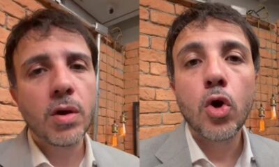Homem com cabelo escuro e barba, vestindo terno claro, fala em frente a uma parede de tijolos